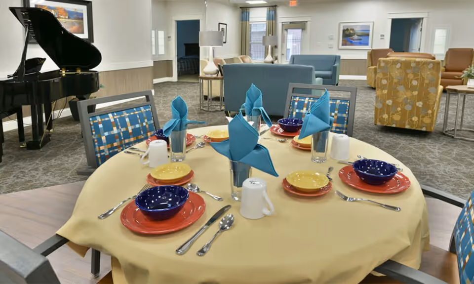 A round dining table set for six with colorful plates, bowls, silverware, and blue folded napkins in glasses. The table is covered with a yellow tablecloth. In the background, there is a black grand piano, several armchairs, sofas, lamps, and framed artwork on the walls in a spacious, carpeted room.