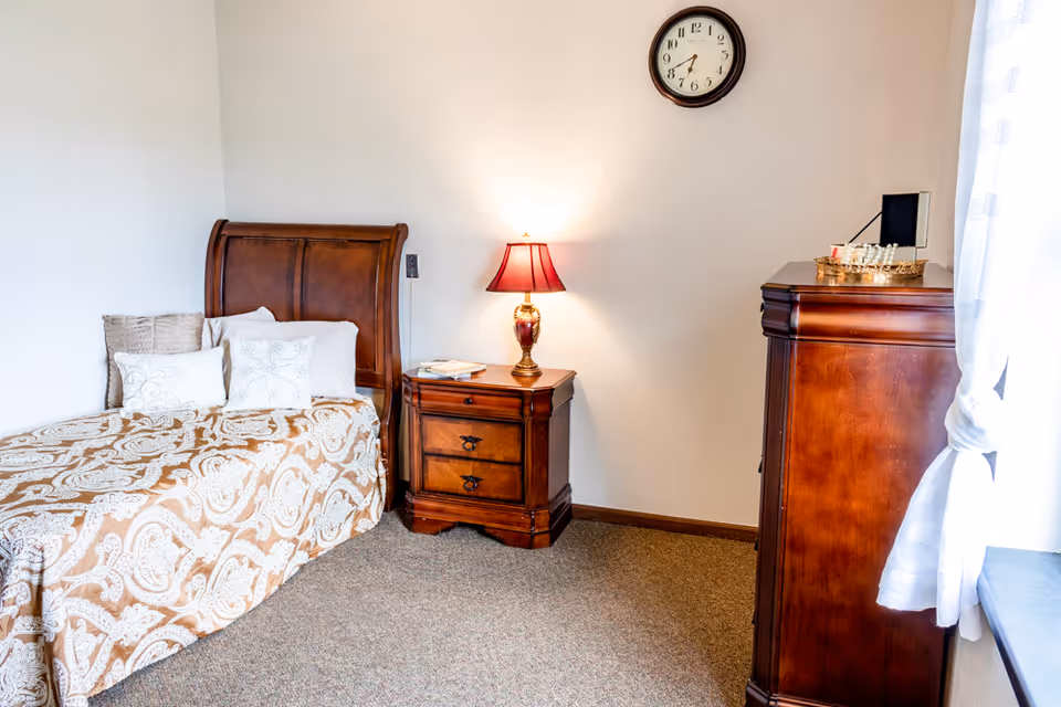 A cozy bedroom with a single wooden bed featuring a patterned bedspread and several pillows. Next to the bed is a wooden nightstand with a red lamp and some books. A wooden dresser is positioned near a window with sheer white curtains. A round wall clock hangs above the nightstand on a plain white wall.