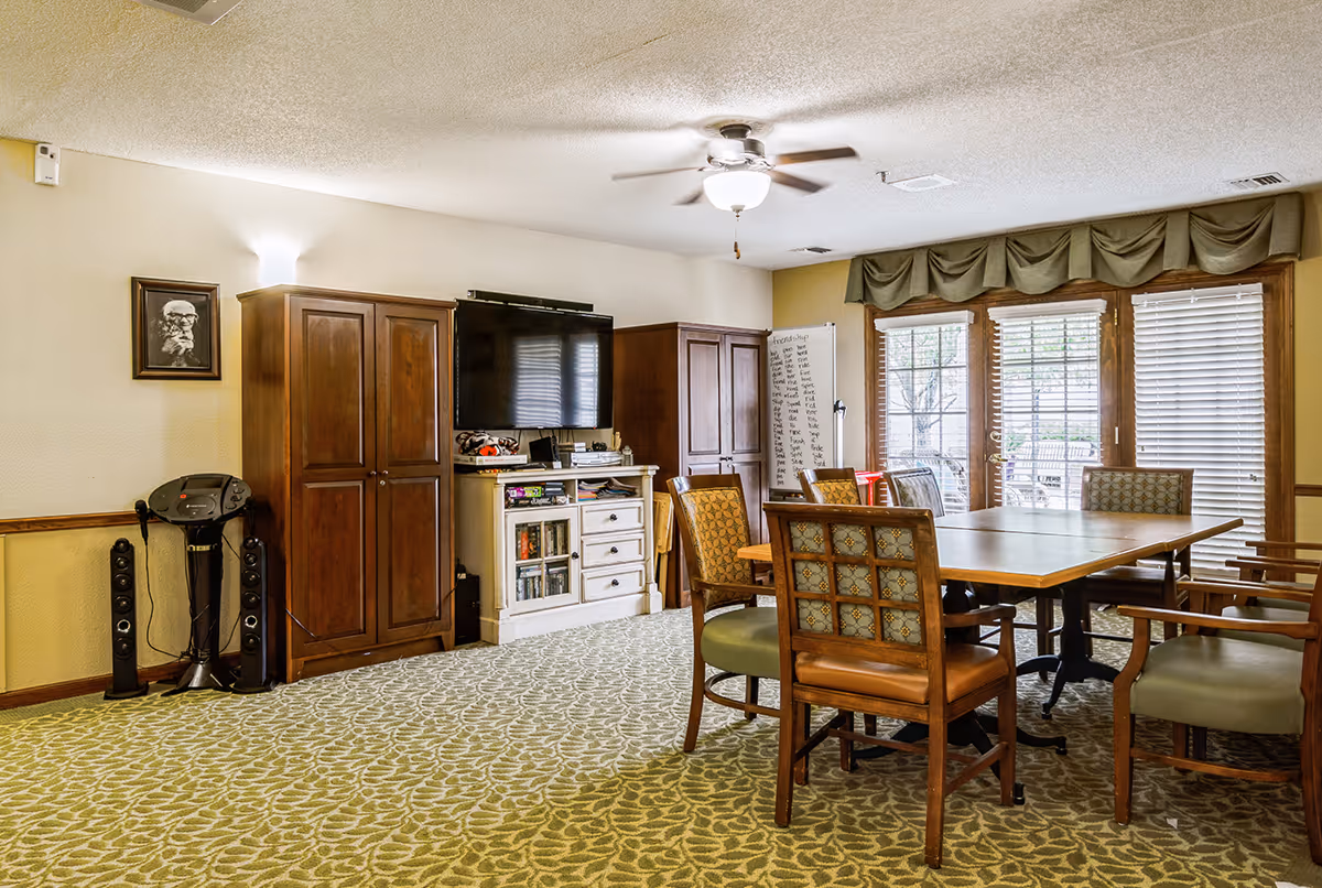 A well-lit common room with a ceiling fan, a large flat-screen TV mounted on the wall, wooden cabinets, a whiteboard with writing, and a table surrounded by six chairs. The room has patterned carpet flooring and large windows with blinds and green valances, allowing natural light to enter.