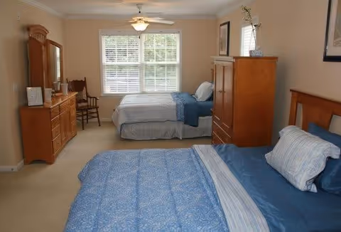 A bedroom with two beds, each made with blue and white bedding. The room has beige walls and carpet, a wooden dresser with a mirror, a wooden chest of drawers, a wooden chair, and a ceiling fan with a light. A window with white blinds lets in natural light.