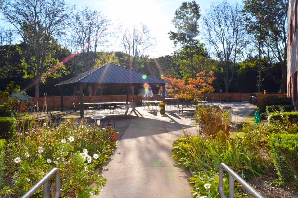 Sunlit outdoor courtyard with a central gazebo, tables and chairs, and plant beds flanking a concrete walkway.