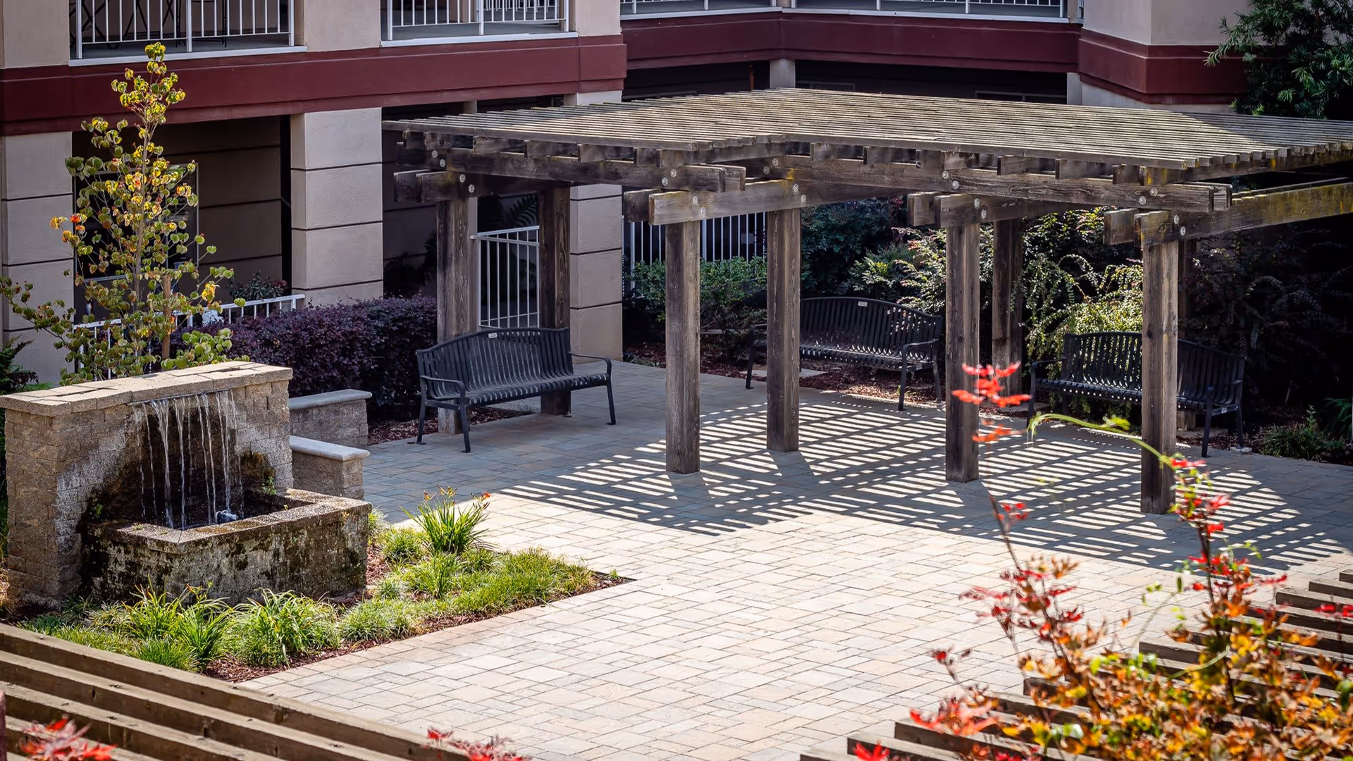 Outdoor courtyard area with a wooden pergola casting shadows on a paved patio. There are three black metal benches under the pergola and a stone water fountain with water flowing down into a basin. Surrounding the patio are various plants and shrubs, with a building facade in the background.
