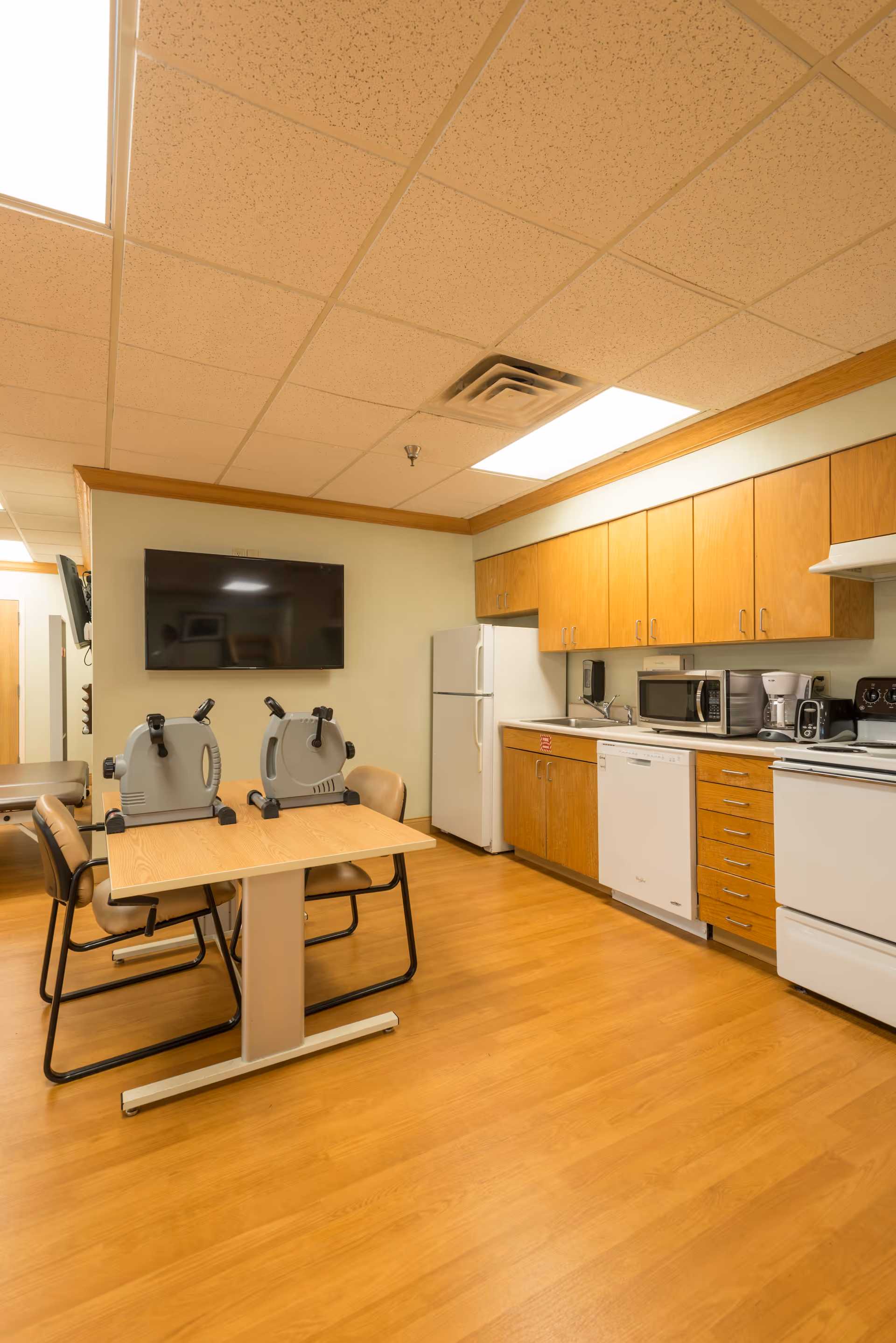 A kitchen area in a senior living facility with wooden cabinets, a white refrigerator, dishwasher, microwave, coffee maker, and stove. There is a wooden table with two small exercise bikes on it and four chairs around the table. A flat-screen TV is mounted on the wall above the table. The floor is wood laminate and the ceiling has recessed lighting.