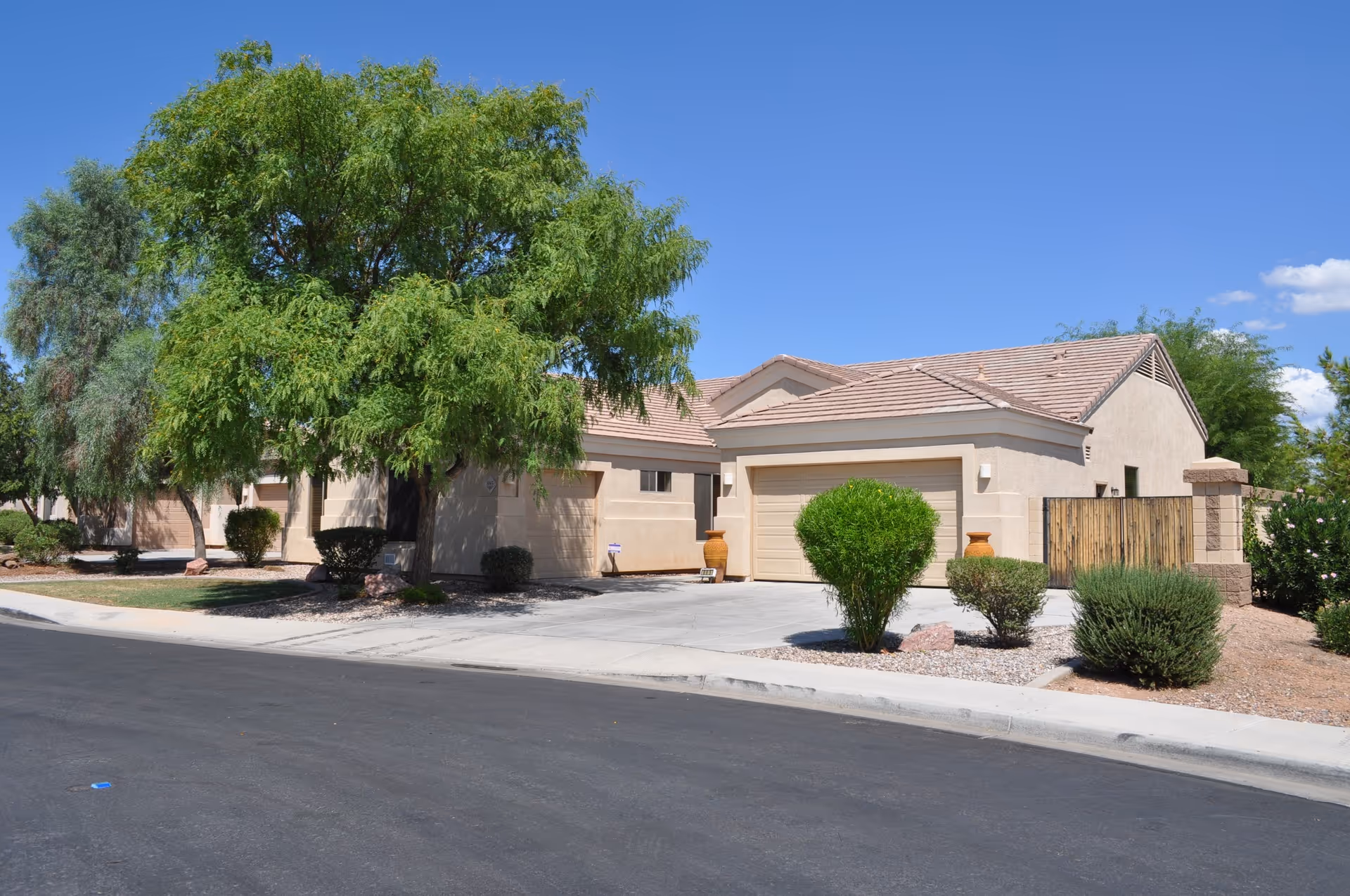 Exterior view of a single-story residential building with a beige facade and tiled roof, surrounded by green trees and shrubs under a clear blue sky.