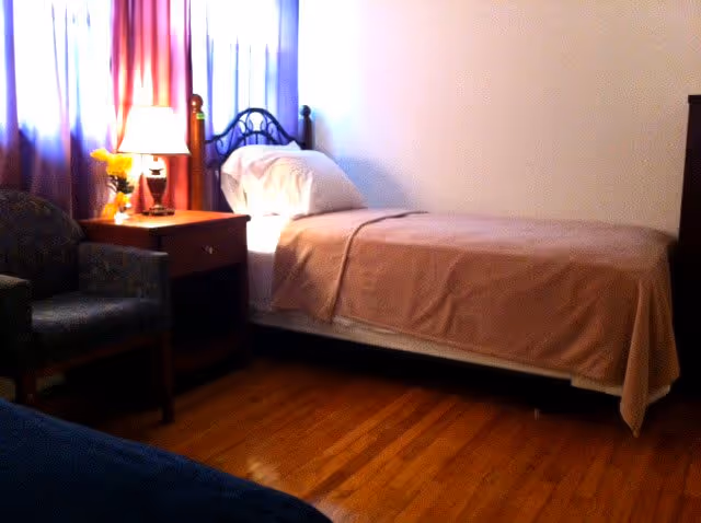 A bedroom with a single bed covered with a light brown blanket and a white pillow. Next to the bed is a wooden nightstand with a lit lamp and a small vase of yellow flowers. There is a cushioned armchair beside the nightstand. The room has wooden flooring and light-colored walls, with windows covered by curtains in shades of red and white.