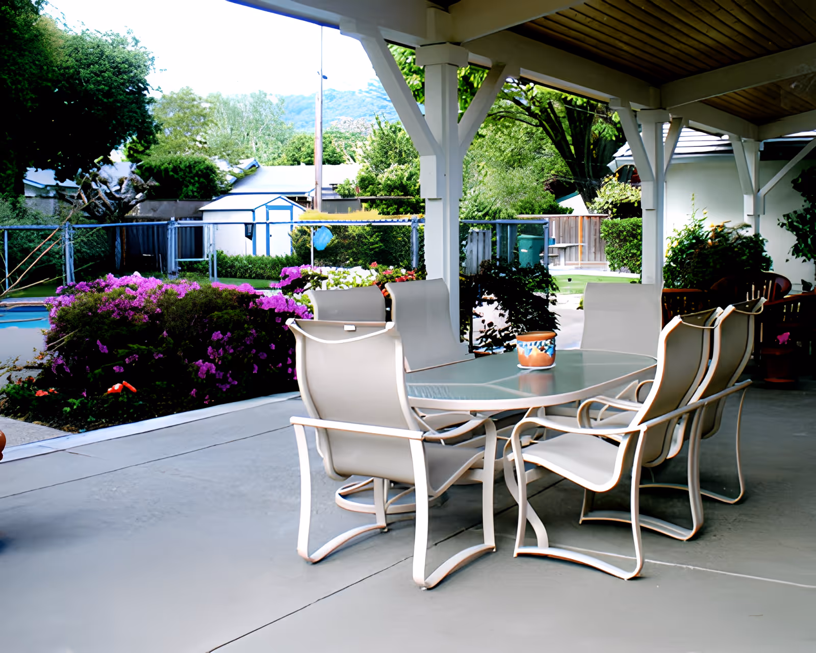 Covered outdoor patio area with a glass-top table and six beige cushioned chairs. The patio overlooks a garden with blooming purple flowers, green bushes, and trees. In the background, there is a fenced yard with a small shed and a mountain view.