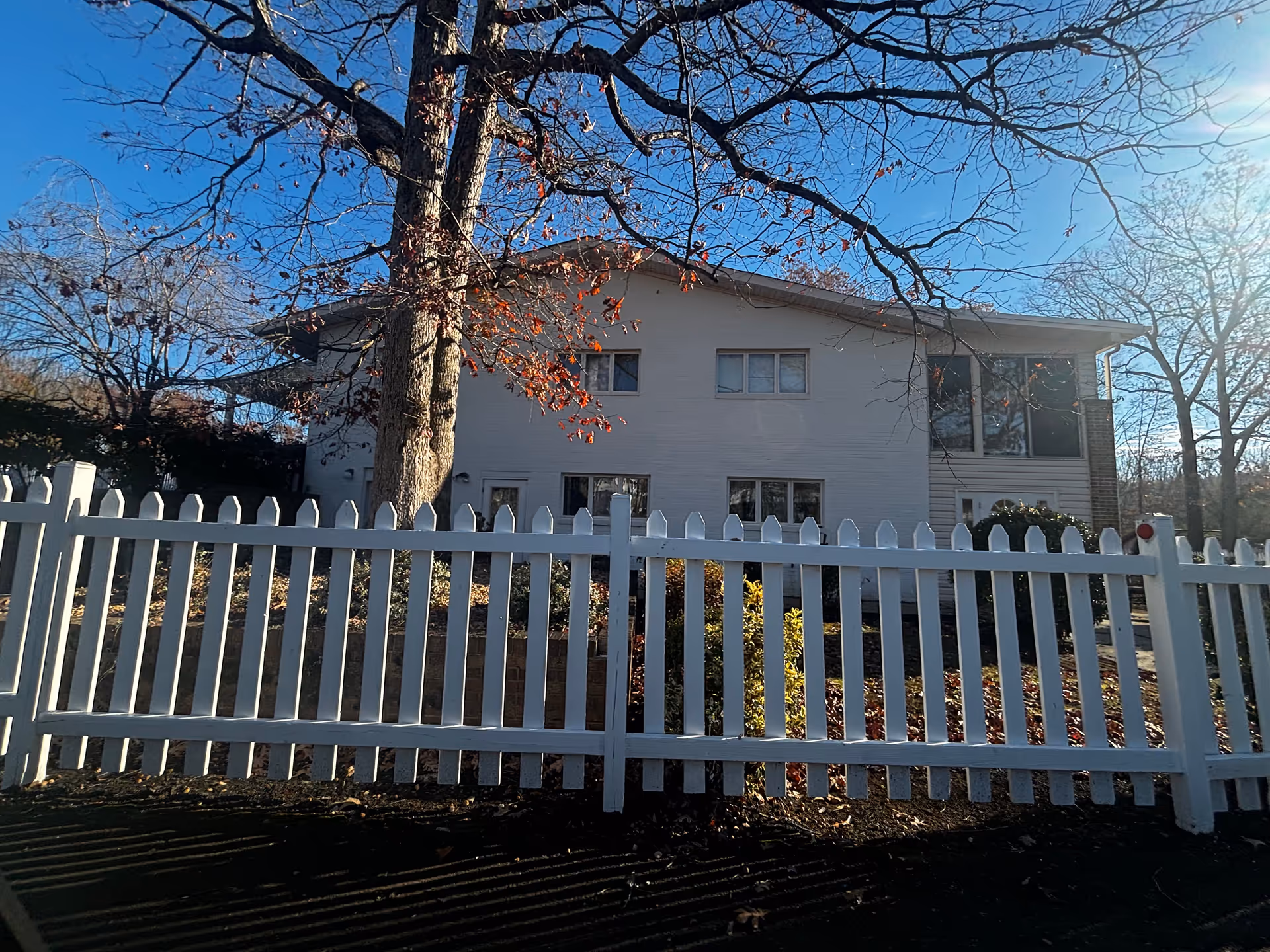 Two-story light-colored building behind a white picket fence with a large tree in front under a clear blue sky.