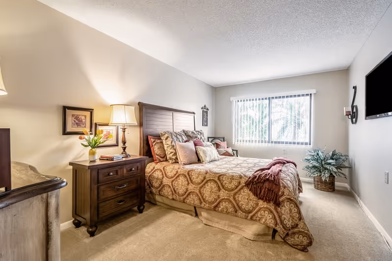 Well-lit furnished bedroom with a double bed dressed in patterned bedding, a nightstand with a lamp and flowers, a wall-mounted TV, and a window with vertical blinds.