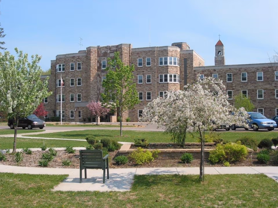 A large brick building with multiple windows and a clock tower in the background, surrounded by a landscaped garden with flowering trees, shrubs, a green bench, and a paved walkway under a clear blue sky.