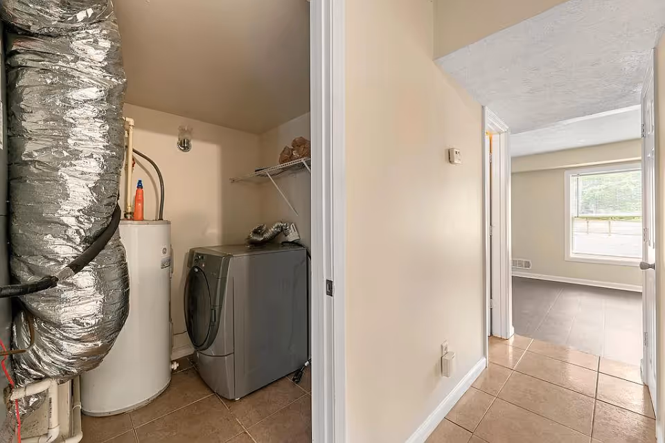 A small laundry room with a washing machine, water heater, and large insulated ductwork. Adjacent to the laundry room is a hallway leading to a room with a window and tiled floor.