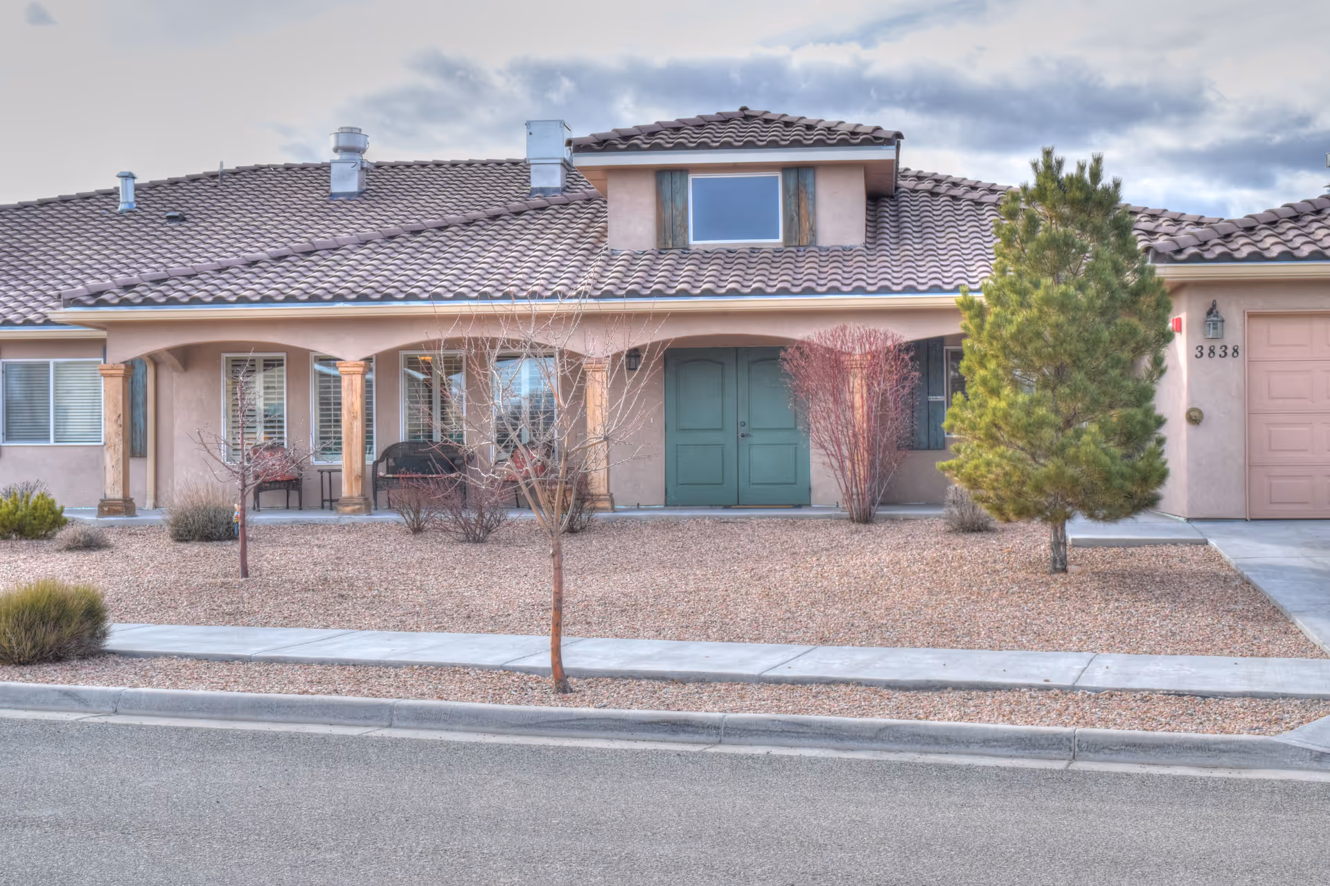 Front exterior view of a single-story residential building with a tiled roof, green double front doors, a small porch with wooden columns, and a gravel yard with sparse vegetation including small trees and bushes.