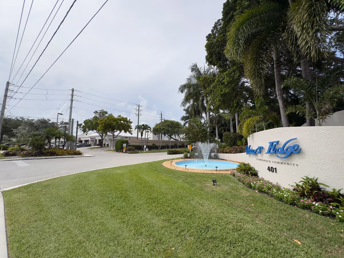 Entrance to Harbour's Edge showing a curved sign, fountain, landscaped lawn, palm trees and driveway.