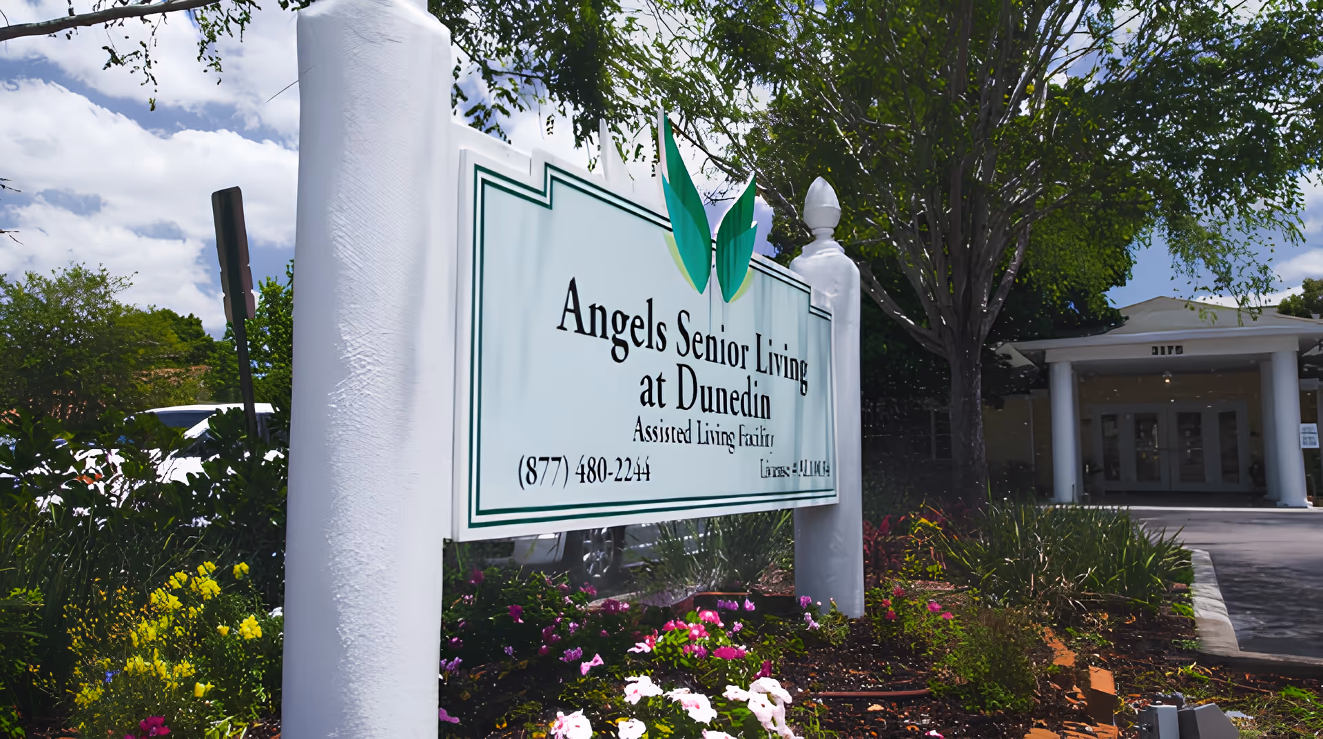 Outdoor view of the entrance to Angels Senior Living at Dunedin, showing a white sign with green accents and text, surrounded by colorful flowers and greenery, with the building entrance and trees in the background under a partly cloudy sky.