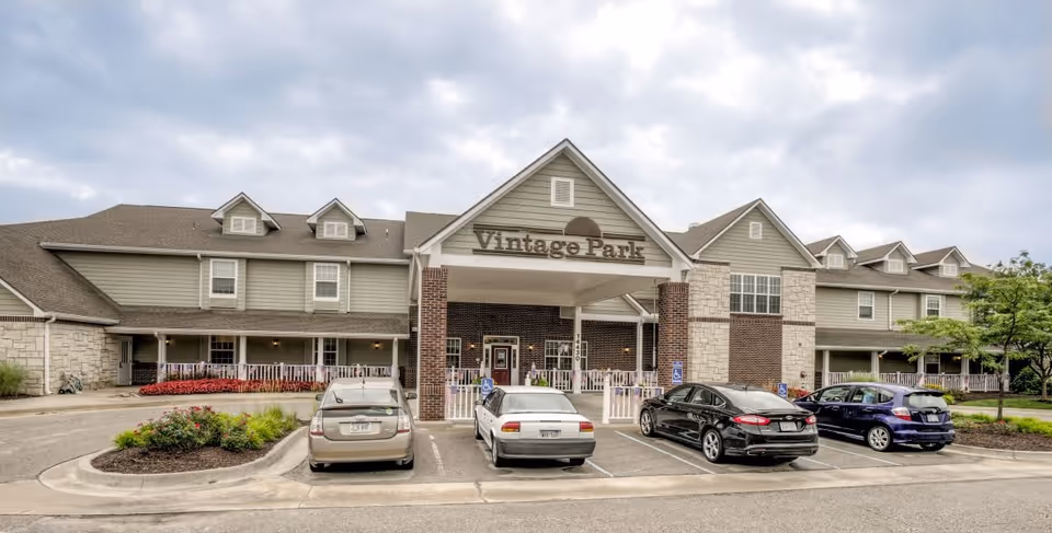 Front exterior view of the Vintage Park at Stanley senior living facility showing a large building with a covered entrance, several parked cars in front, and a cloudy sky above.