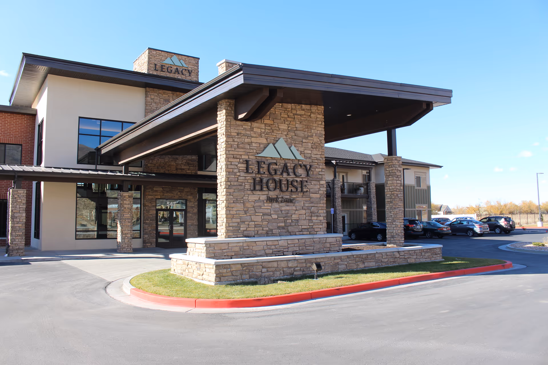 Exterior view of Legacy House of Park Lane building with stone pillars and a covered entrance. Several cars are parked in the parking lot under a clear blue sky.