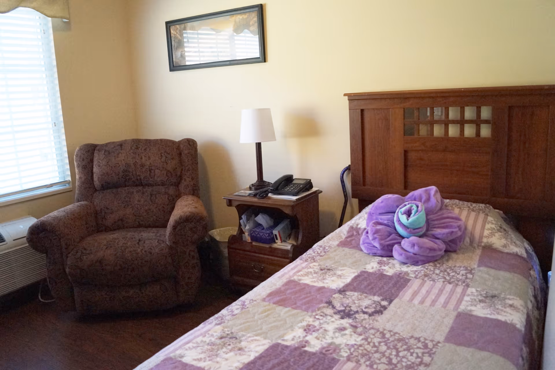 Small bedroom with a single bed with a quilt, wooden headboard, nightstand with a lamp and an upholstered armchair by the window.