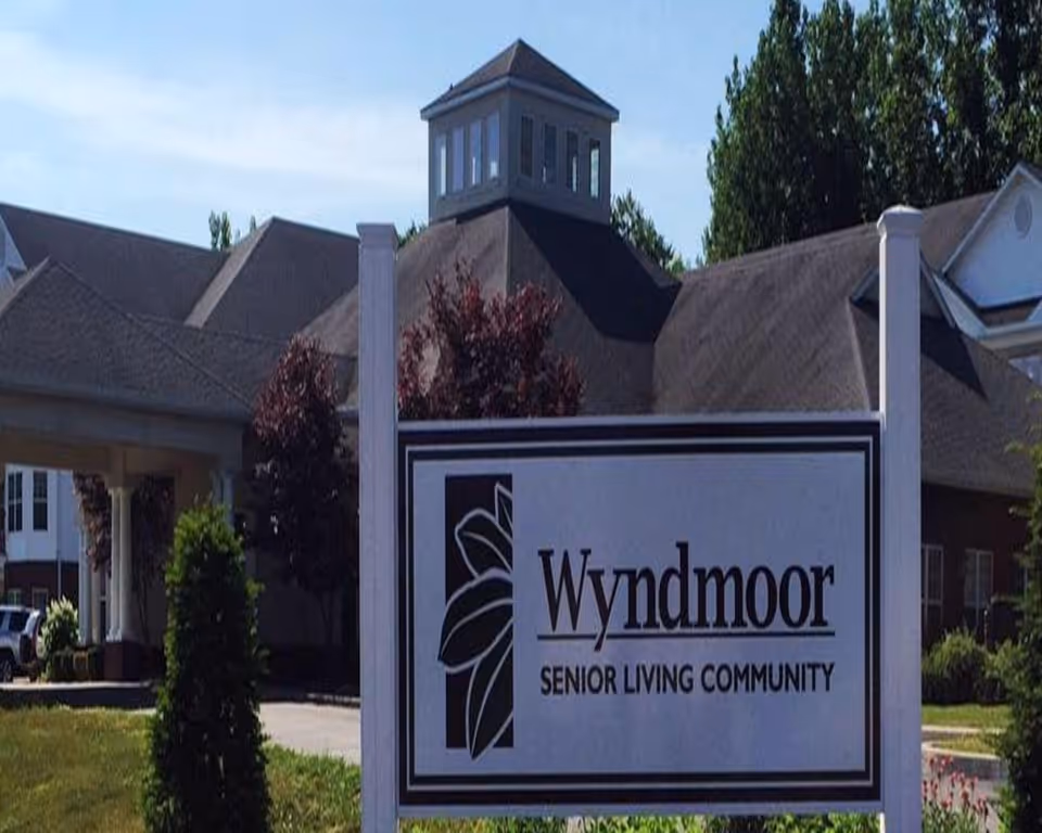 Exterior view of Wyndmoor Senior Living Community building with a large sign in the foreground displaying the facility name and logo. The building has a peaked roof with a cupola and is surrounded by trees and landscaping.