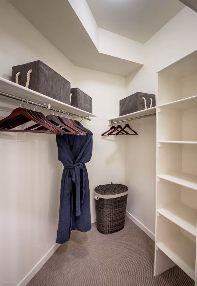 A neatly organized walk-in closet with wooden hangers, gray storage bins on upper shelves, a navy robe hanging, and a wicker laundry basket.