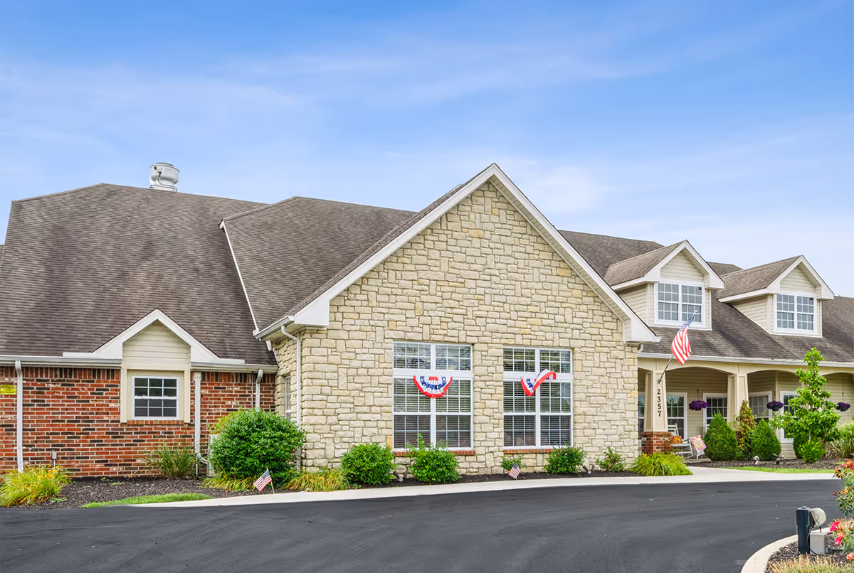Front exterior of a single-story senior living facility with stone and brick facade, American flags, and landscaped driveway.