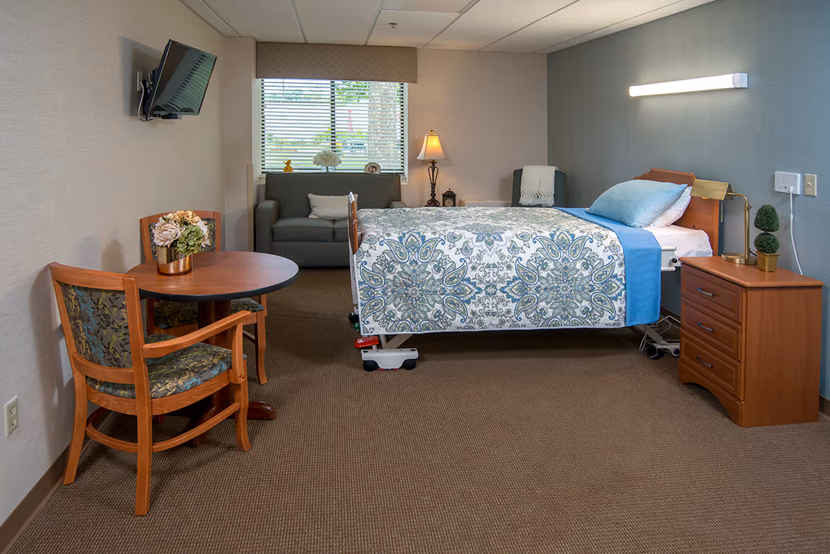 A nursing facility room with a hospital-style bed covered with a patterned blue and white bedspread. Next to the bed is a wooden nightstand with a small lamp and decorative plants. There is a small round wooden table with two wooden chairs featuring floral upholstery. In the background, a window with blinds lets in natural light, and a gray loveseat and a floor lamp are positioned against the wall. A flat-screen TV is mounted on the wall to the left.