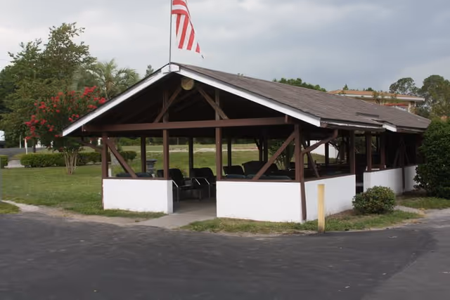 Open-sided pavilion structure with a peaked roof and white lower walls, containing several black chairs inside. An American flag is mounted on top of the roof. The pavilion is situated on a paved area with grass and trees in the background under a cloudy sky.