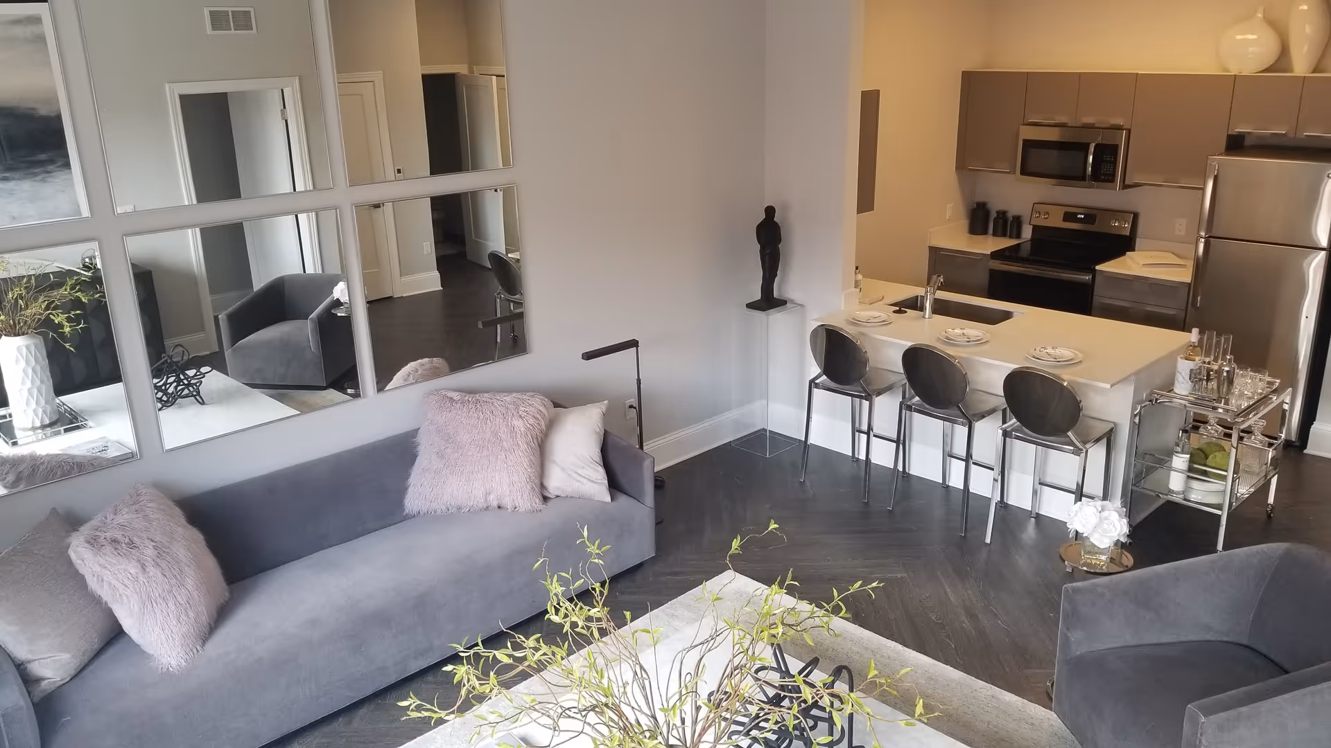 Modern open-concept living area with a gray sofa adorned with pink and gray pillows, a matching gray armchair, a white rug with a plant centerpiece, and a kitchen area with a breakfast bar featuring three black bar stools. The kitchen includes stainless steel appliances and light-colored cabinetry. Large square mirrors are mounted on the wall above the sofa, reflecting parts of the room.