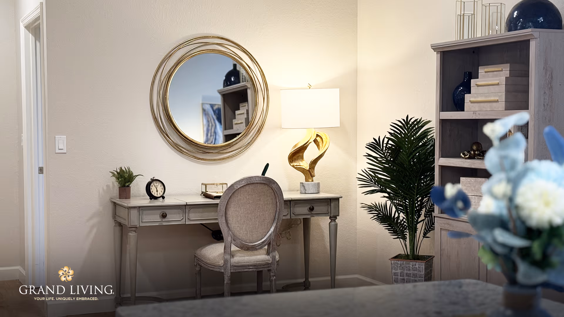 A cozy interior corner featuring a vintage-style desk with a round mirror above it, a decorative gold lamp, a small clock, and a potted plant. To the right, there is a tall wooden bookshelf with decorative items and a large green plant beside it. In the foreground, a blurred bouquet of flowers is partially visible.