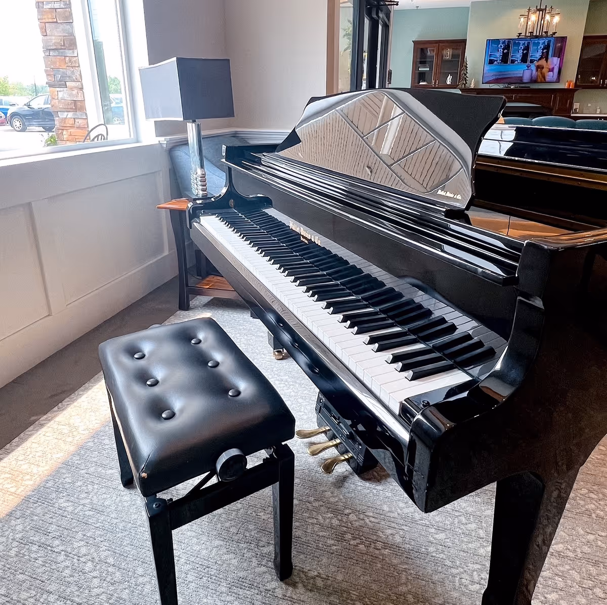 A black grand piano with a matching black cushioned bench in a well-lit room. The piano is positioned near a window with natural light coming in. In the background, there is a lamp on a small table and a television mounted on the wall above a wooden cabinet.