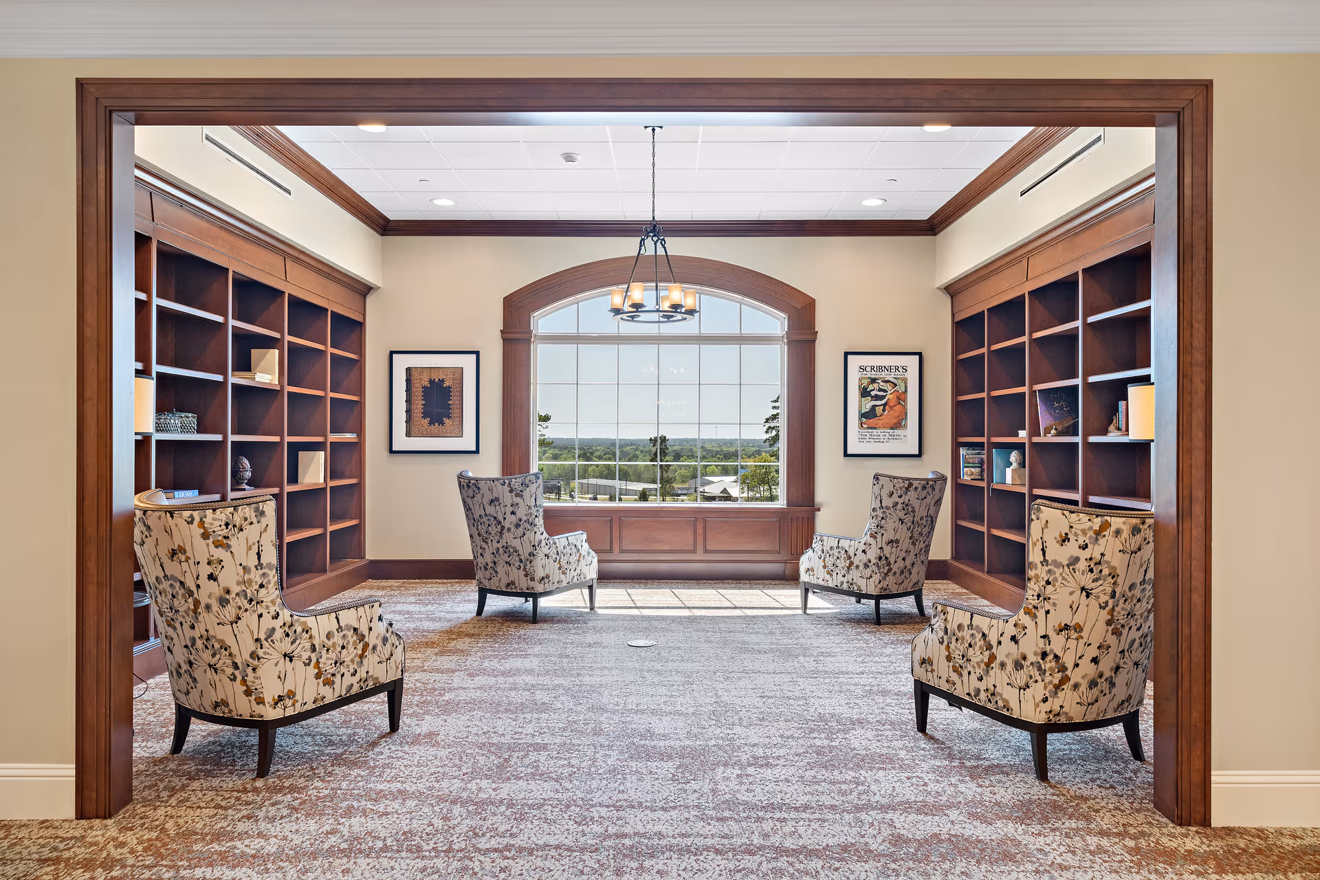 A cozy reading or sitting room with four floral-patterned armchairs arranged in pairs facing each other. The room features large wooden bookshelves on both sides, a large arched window at the far end letting in natural light, and framed artwork on the walls. The carpet has a soft, patterned design and a chandelier hangs from the ceiling.