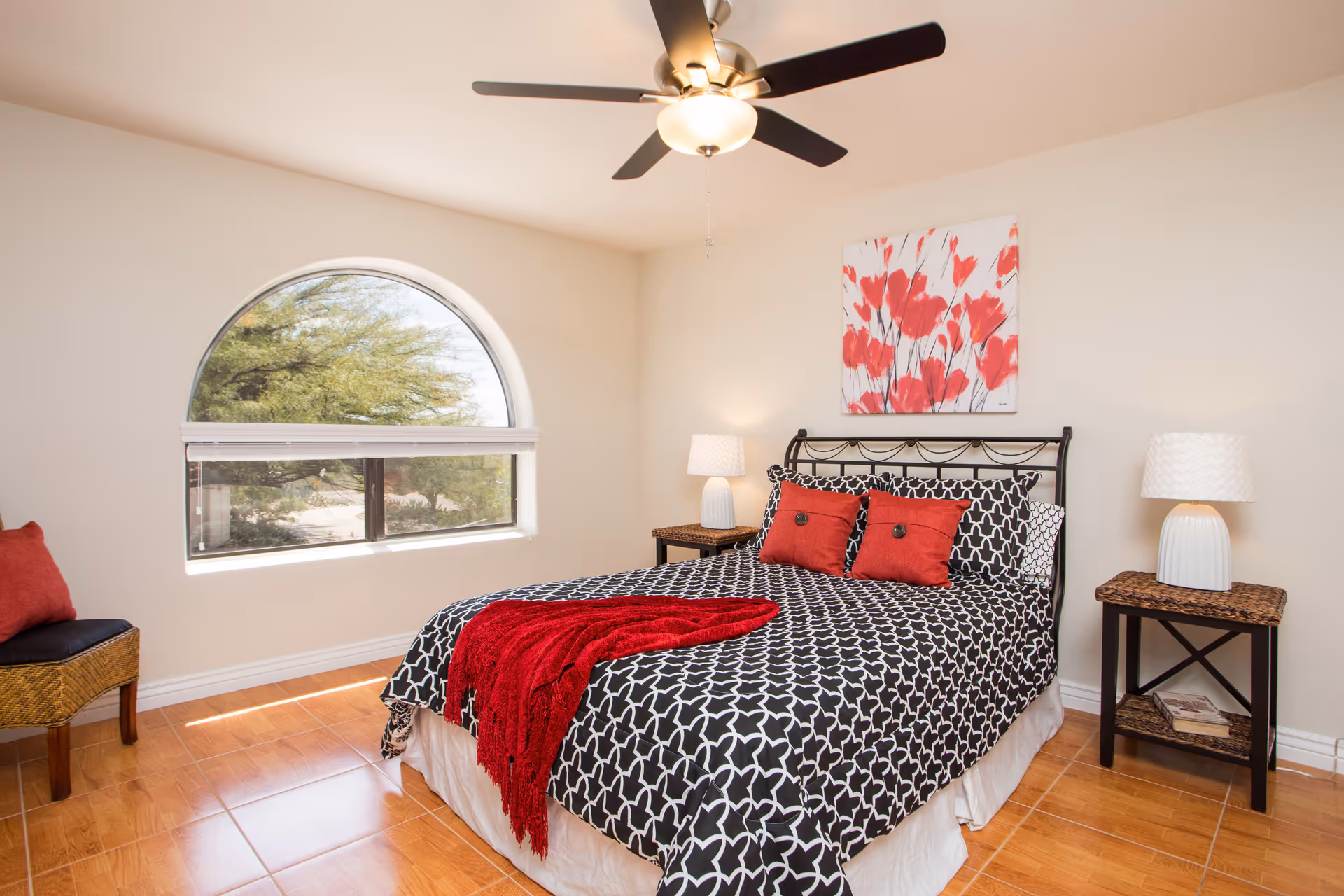 A bright bedroom with a large arched window showing trees outside. The room features a bed with black and white patterned bedding, two red pillows, and a red throw blanket. There are two wicker nightstands with white lamps on either side of the bed, and a ceiling fan with a light above. A painting with red flowers hangs on the wall above the bed.