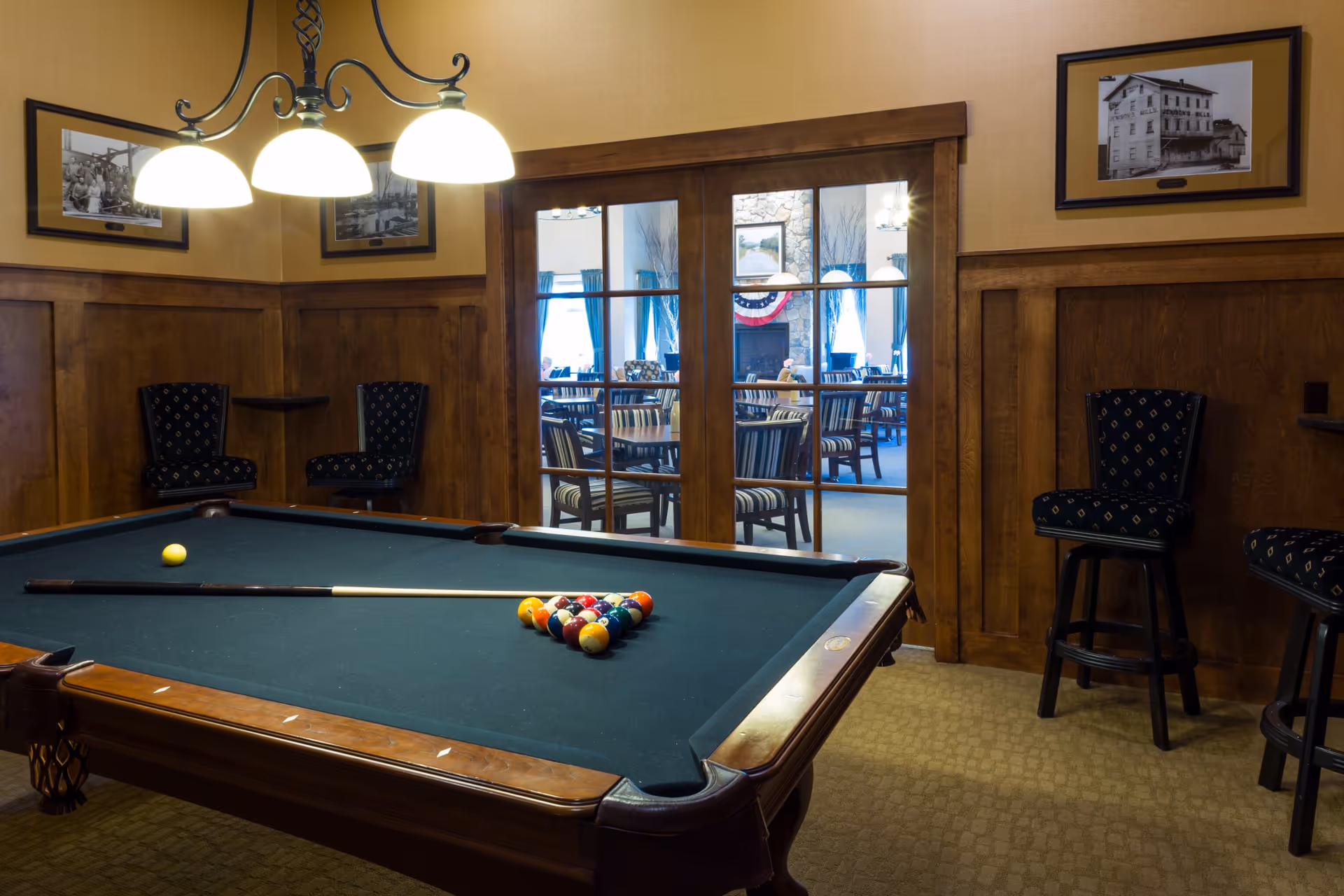 Wood-paneled interior game room with a pool table, cue stick and seating, and glass doors opening to a dining area.