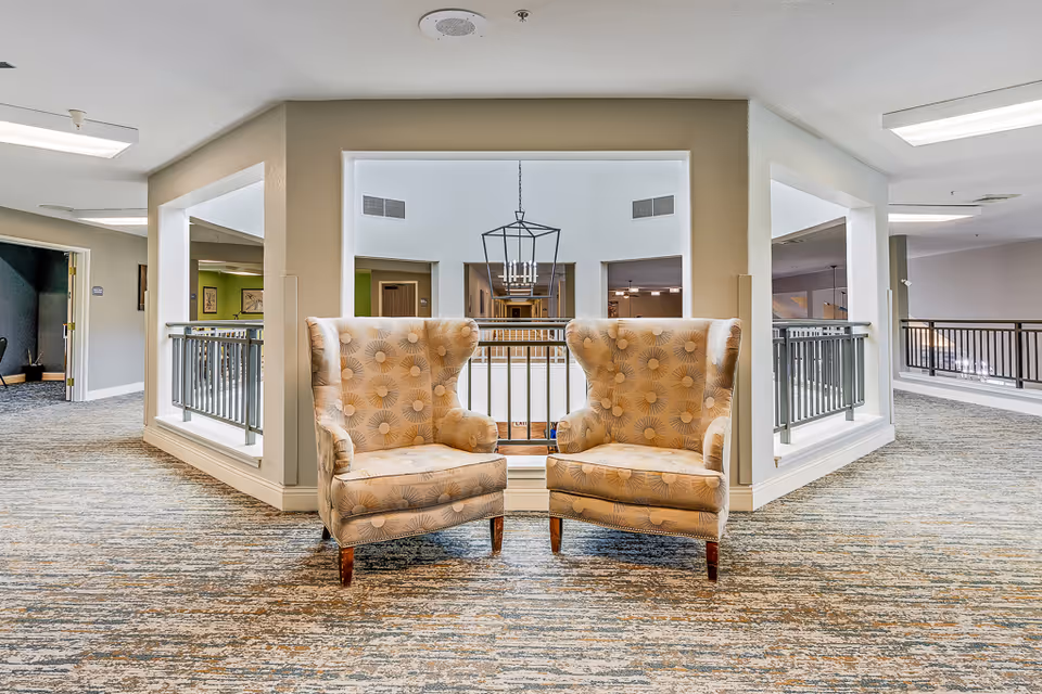 Two patterned beige armchairs placed side by side in a carpeted hallway area with railings overlooking a lower floor. The space is well-lit with ceiling lights and has neutral-colored walls and carpet.
