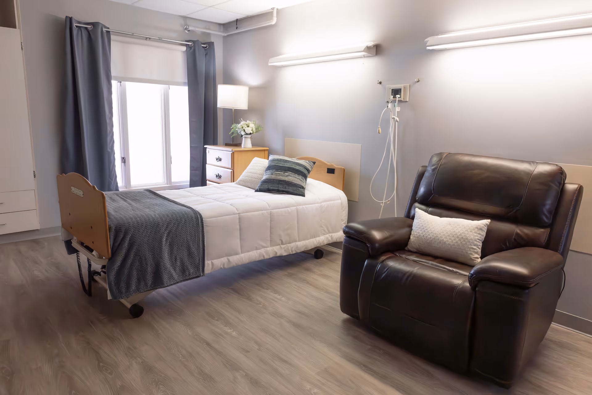 A neatly arranged senior living bedroom featuring a single bed with white bedding and a gray throw blanket, a wooden dresser with a vase of flowers, a large window with gray curtains, and a dark brown leather recliner chair with a white cushion. The room has light gray walls and wood-patterned flooring.