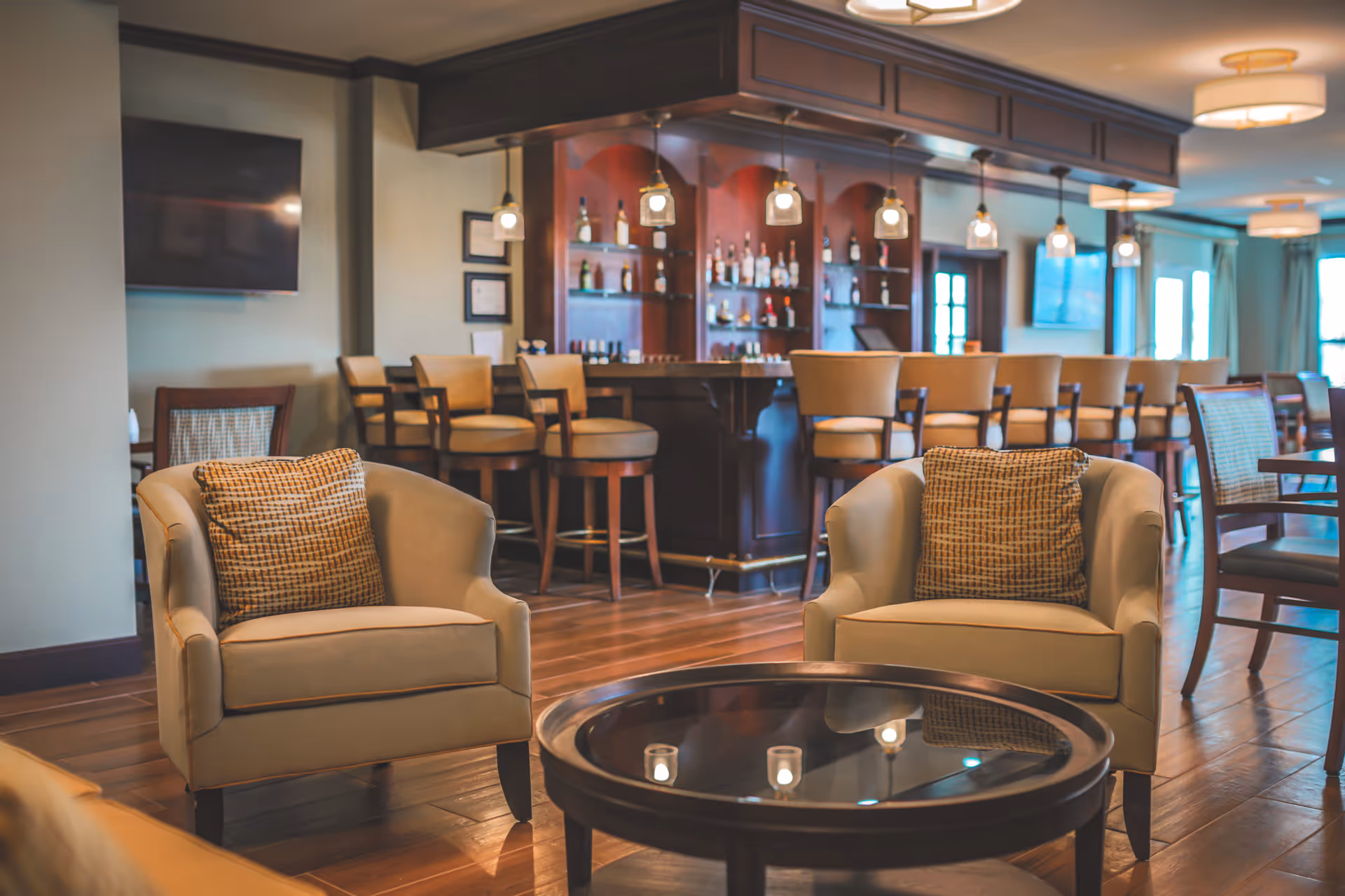 A cozy lounge area with two beige armchairs with patterned cushions facing a round glass coffee table. In the background, there is a wooden bar with several bar stools and shelves stocked with bottles. The room has warm lighting and wooden flooring.