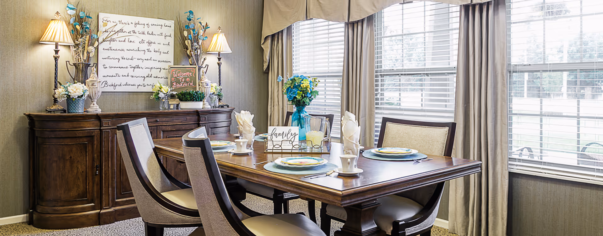 A well-lit dining room with a wooden dining table set for four, featuring plates, cups, and napkins. The table has a centerpiece with a vase of blue and yellow flowers and a sign that says 'family'. Behind the table is a wooden sideboard decorated with lamps, flower arrangements, and a small chalkboard sign that reads 'Welcome to Bickford'. Large windows with beige curtains allow natural light to fill the room.
