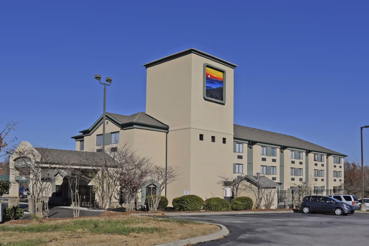 Front exterior of a three-story senior living building with a central tower, entrance canopy, landscaping, and a parking lot under a clear blue sky.