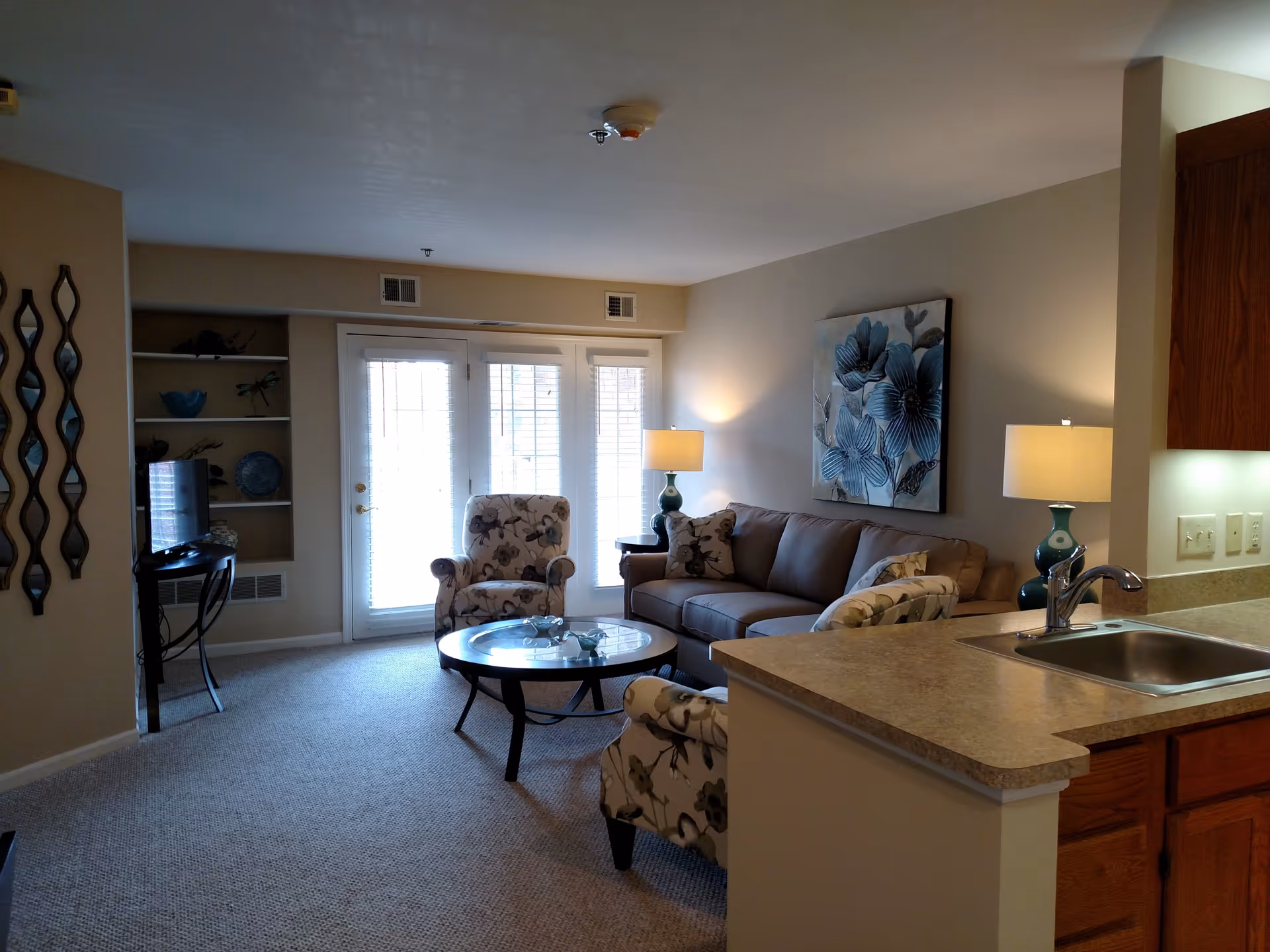 A cozy living room area with a beige sofa, two floral patterned armchairs, a round glass coffee table, and two table lamps with blue bases. There is a large floral painting on the wall above the sofa. To the right, a kitchen counter with a sink is visible. French doors with blinds lead outside, and a small TV is placed on a stand in a built-in shelving nook on the left side of the room.