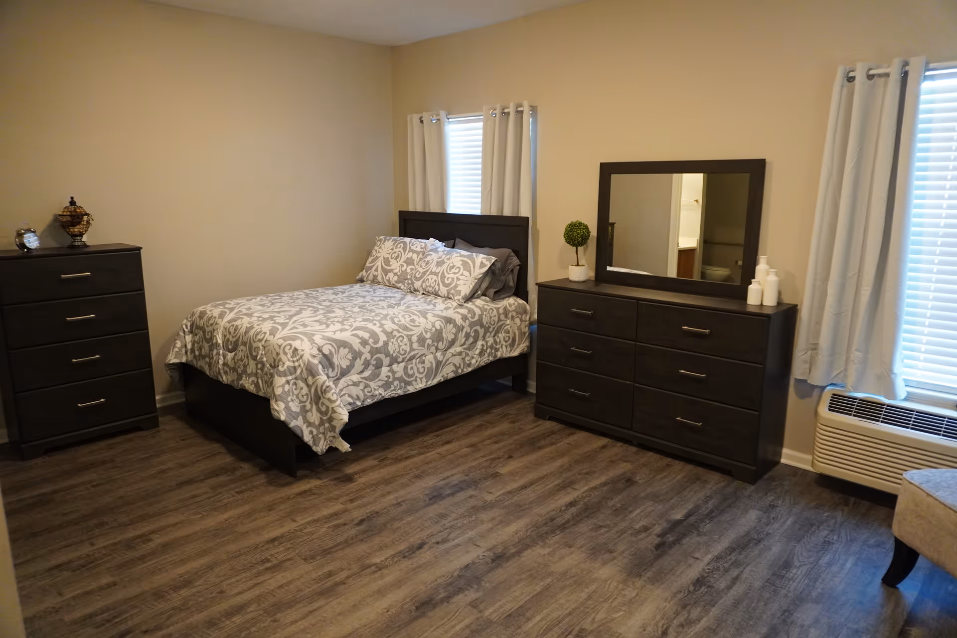 A bedroom with a bed covered in a gray and white patterned comforter, two pillows, a dark wooden headboard, a matching dresser with a mirror, a chest of drawers, two windows with white curtains, and a small potted plant on the dresser. The floor is wood, and there is an air conditioning unit under one window.