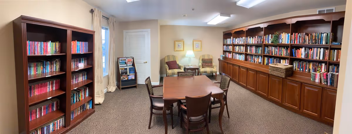 A cozy library room with two large wooden bookshelves filled with books on the left and right walls. In the center, there is a wooden table surrounded by six chairs. At the back of the room, two upholstered armchairs with a small table and lamp between them are positioned against a beige wall with two framed pictures. A window with curtains is on the left side, and a small magazine rack is near the door.