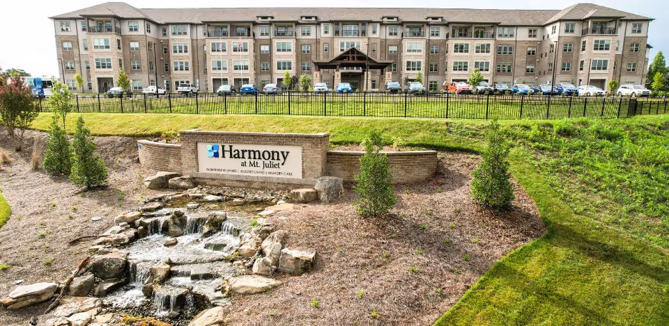 Exterior view of Harmony at Mt. Juliet senior living facility showing a multi-story building with numerous windows and a parking area in front. In the foreground, there is a landscaped area with a small cascading water feature and a sign that reads 'Harmony at Mt. Juliet' along with services offered: Independent Living, Assisted Living, Memory Care.