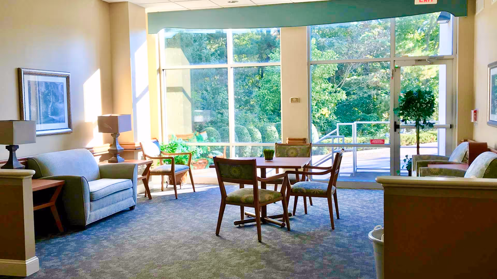 A bright and cozy sitting area with a small square table surrounded by four chairs with patterned cushions. There are two armchairs and a loveseat along the walls, each accompanied by a side table with a lamp. Large floor-to-ceiling windows and a glass door provide a view of lush green trees and bushes outside. The room has carpeted flooring and framed artwork on the walls.