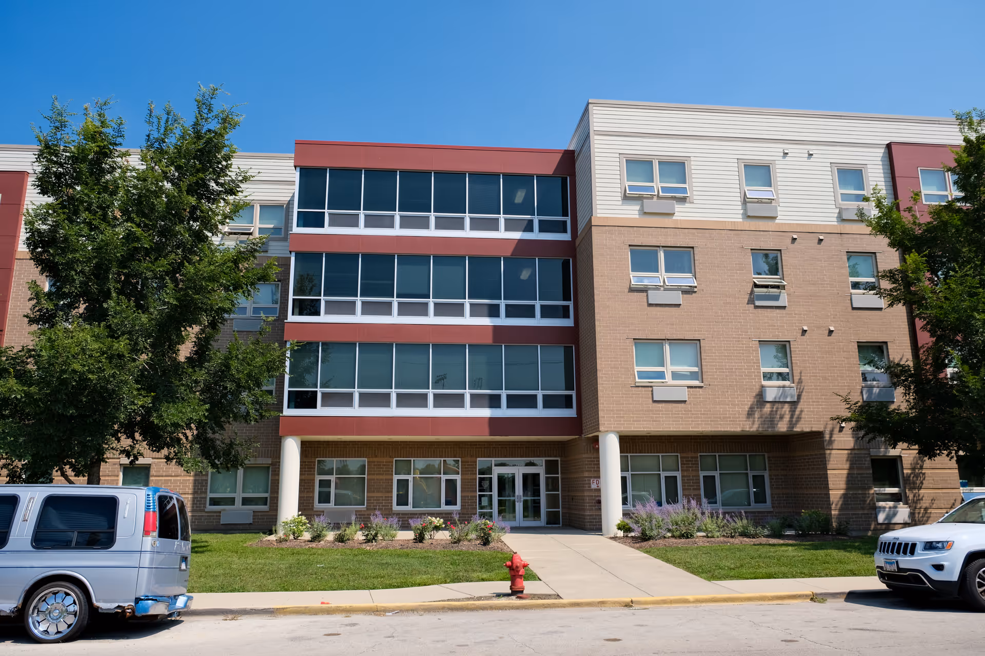 Exterior front view of a modern three-story senior living facility building with large windows, brick and siding facade, a sidewalk leading to the entrance, green lawn, trees, and parked vehicles on the street.