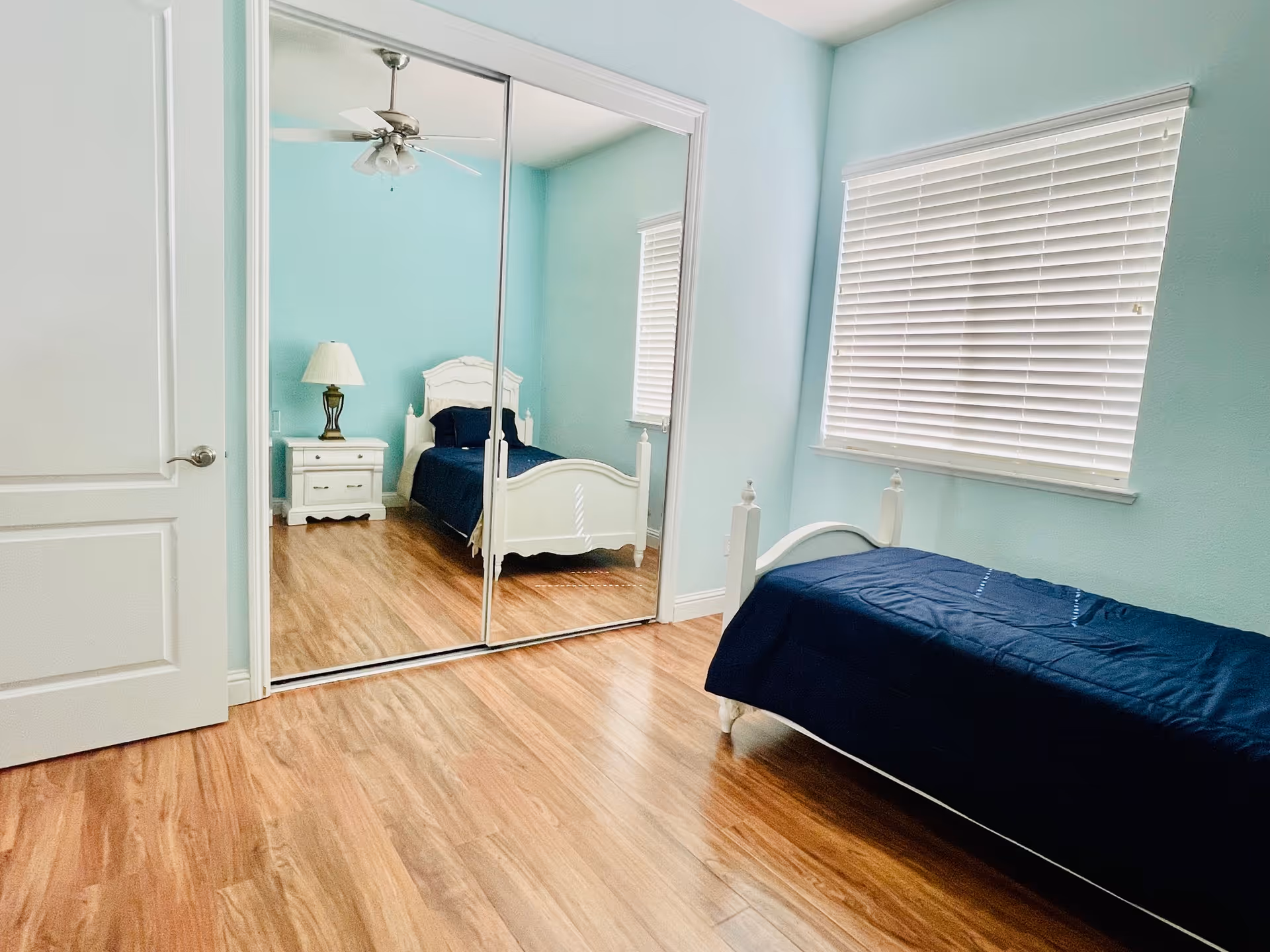 A bedroom with light blue walls, a wooden floor, a white single bed with a dark blue bedspread, a white nightstand with a lamp, a ceiling fan, and a large mirrored closet door reflecting the bed and nightstand.