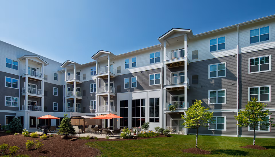 Exterior courtyard of a four-story senior living building with balconies, patio seating and orange umbrellas.