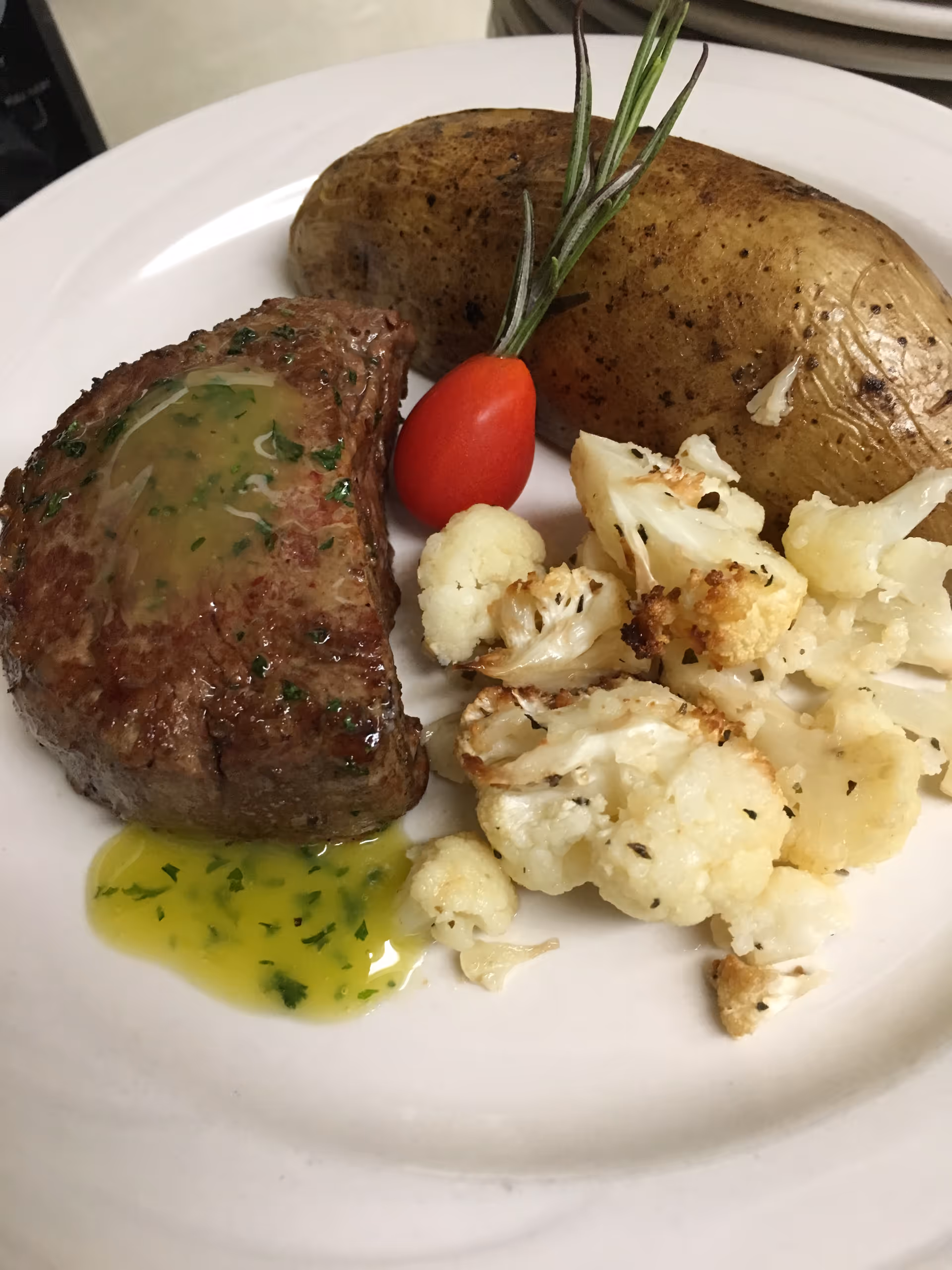 A plated meal consisting of a cooked steak topped with herb butter, a baked potato garnished with a sprig of rosemary and a small red pepper, and roasted cauliflower florets.