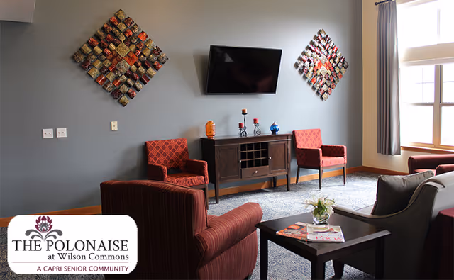 A cozy living room area with a gray wall featuring two decorative square wall art pieces and a flat-screen TV mounted between them. Below the TV is a wooden cabinet with decorative items on top. The room has two red patterned armchairs, a striped armchair, a gray armchair, and a coffee table with magazines and a small flower arrangement. A large window with gray curtains allows natural light into the room.