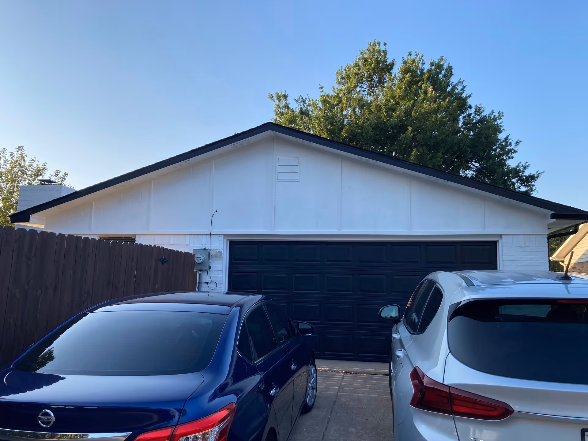 View of a residential garage with a black garage door, flanked by a wooden fence on the left. Two cars, one blue and one silver, are parked in the driveway in front of the garage. There are trees visible behind the garage under a clear blue sky.