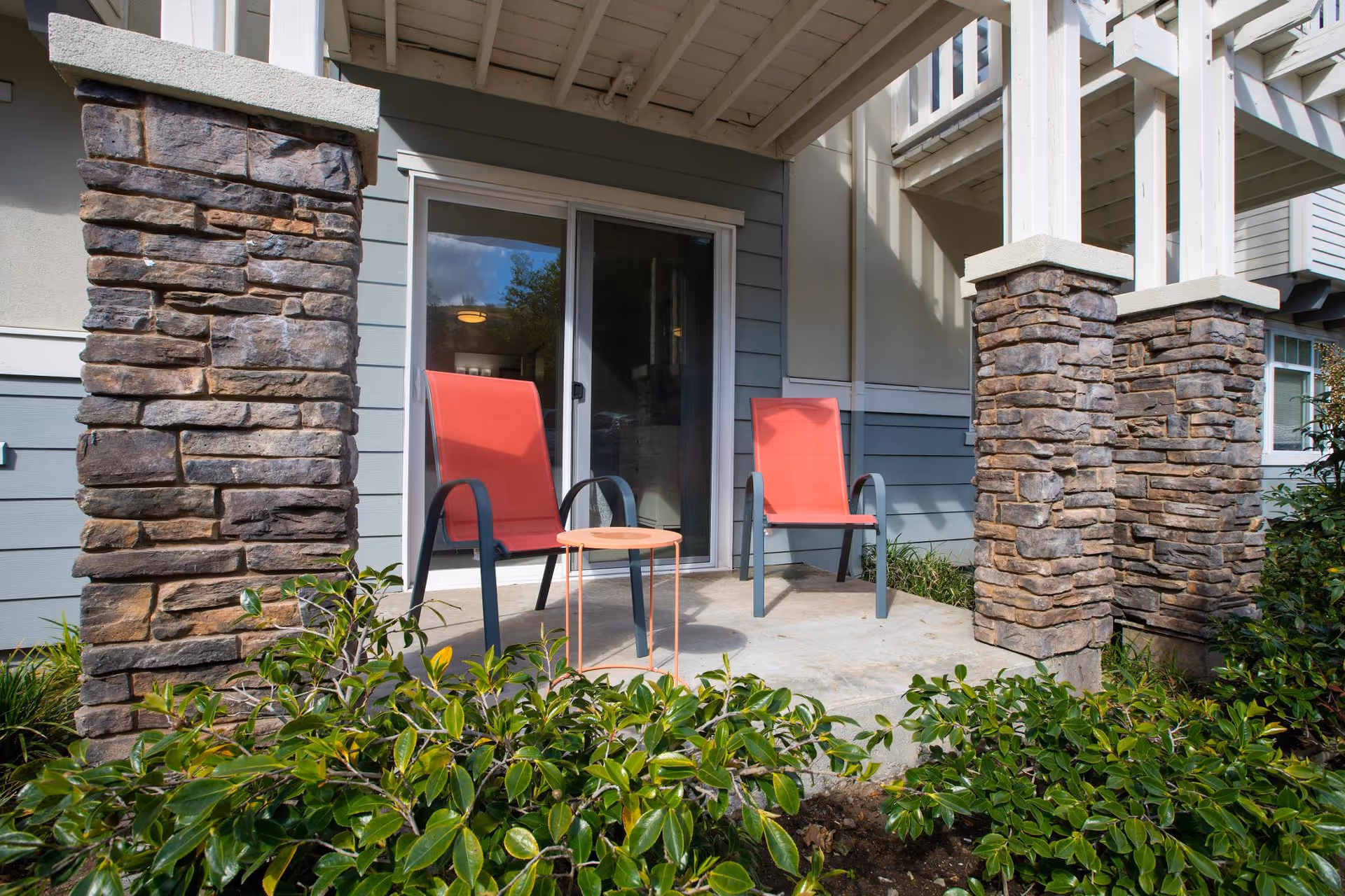 Outdoor patio area with two red chairs and a small round table between them, surrounded by stone pillars and green shrubbery in front of a sliding glass door.