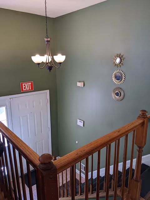 Interior view of a stairwell with wooden handrails and green walls. A white door with an exit sign above it is visible at the bottom of the stairs. Three decorative sunburst mirrors are mounted on the wall, and a chandelier with five lights hangs from the ceiling.