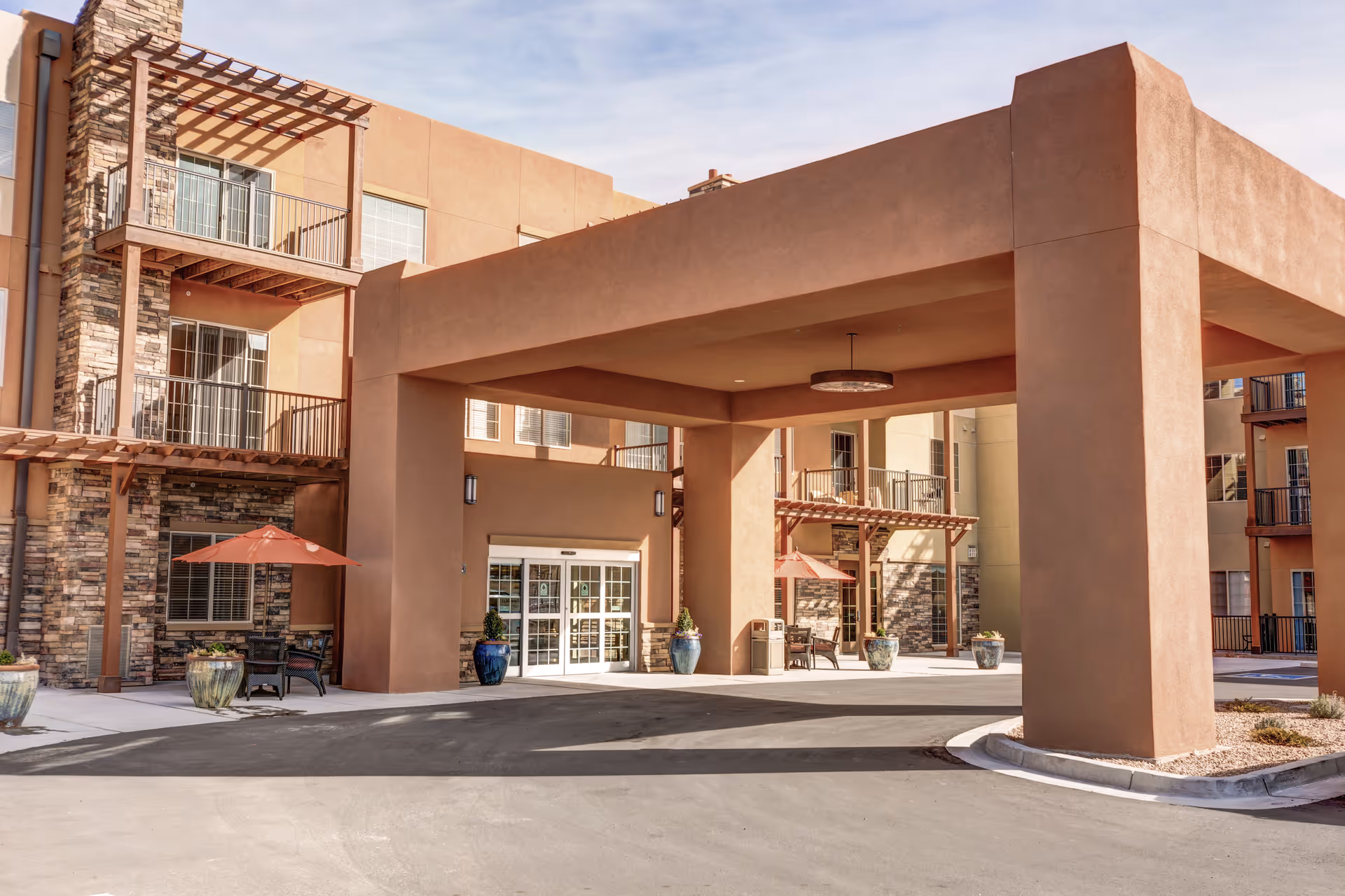 Exterior view of the entrance to a senior living facility with a large covered drop-off area supported by thick columns. The building features a southwestern architectural style with stucco walls and stone accents. There are balconies with metal railings and outdoor seating areas with umbrellas near the entrance.